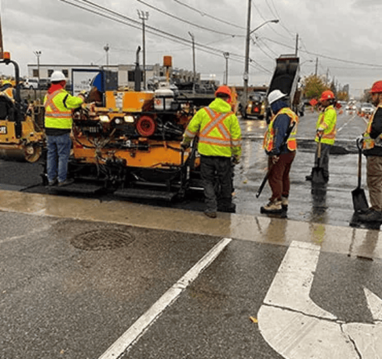 Construction workers paving a road with heavy machinery in wet weather.
