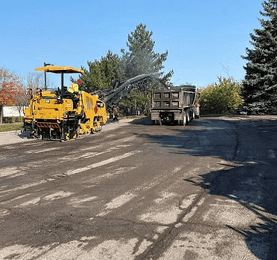 Paving machine working on road construction site with dump truck in the background.