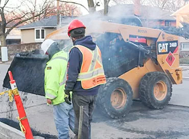 Construction workers operating a skid steer loader on asphalt paving site.